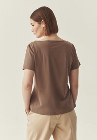 Haut marron à manches courtes avec un large décolleté et une texture lisse. Associé à un pantalon de couleur claire, mettant en valeur une coupe décontractée.