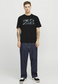 Musta puuvillapaita, jossa on monimallinen "JACK & JONES" -logo. Yhdistetty löysiin sinisiin farkkuihin ja mustiin lenkkareihin.
