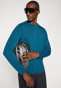 Langärmliges, türkisfarbenes Sportshirt mit flachen Nähten, kombiniert mit einem matten grauen Fahrradhelm. Modell trägt große schwarze Sonnenbrillen.