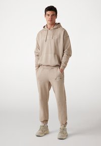 Beige kapucnis pulóver logóval, hozzáillő joggingnadrággal, oldalcsíkkal és rugalmas mandzsettával. Simább anyag, laza szabású.
