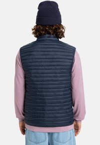 Marineblaue Steppweste mit horizontalem Steppmuster, ausgestattet mit einem hohen Kragen und leichter Textur, über einem hellrosa Langarmshirt getragen.