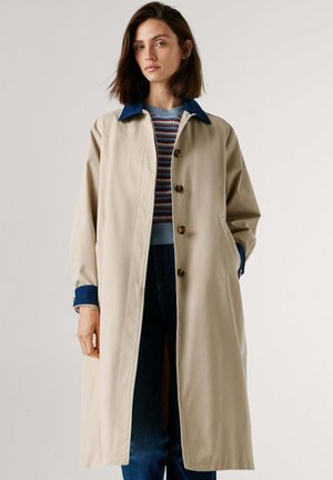 Smėlio spalvos tranšėjos paltas su mėlynu apykakliu, dideliais rudais mygtukais, šoninėmis kišenėmis ir laisvo silueto. Dėvimas ant dryžuoto megztinio.