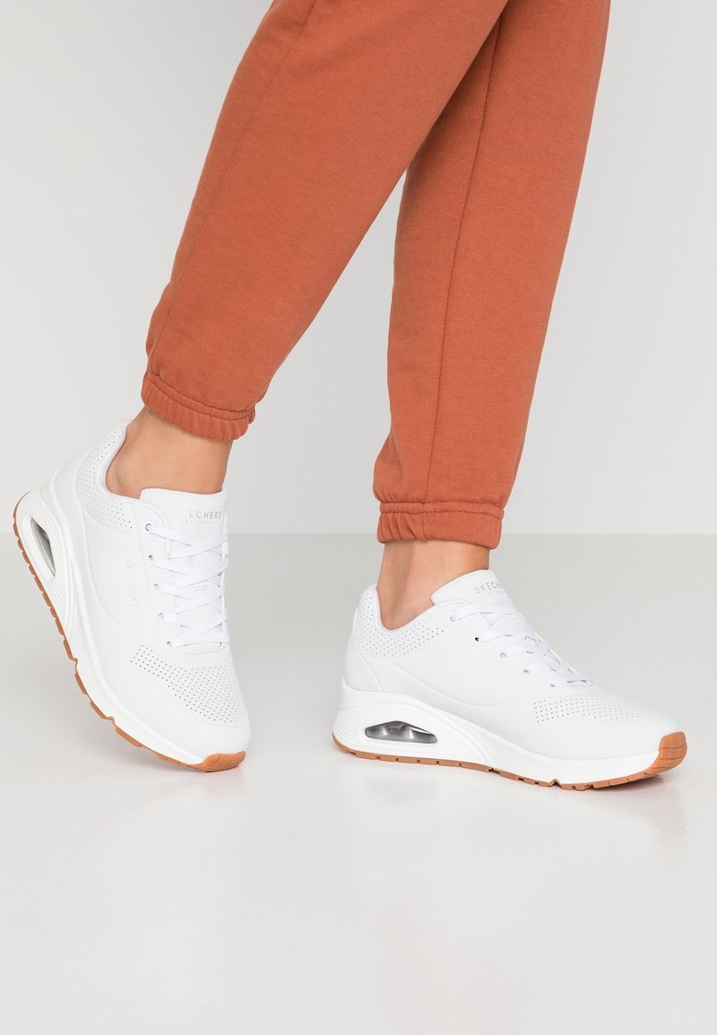 Chaussures de sport blanches avec un design perforé, dotées d'une semelle coussinée et d'une unité d'air transparente, associées à un pantalon de survêtement couleur rouille.