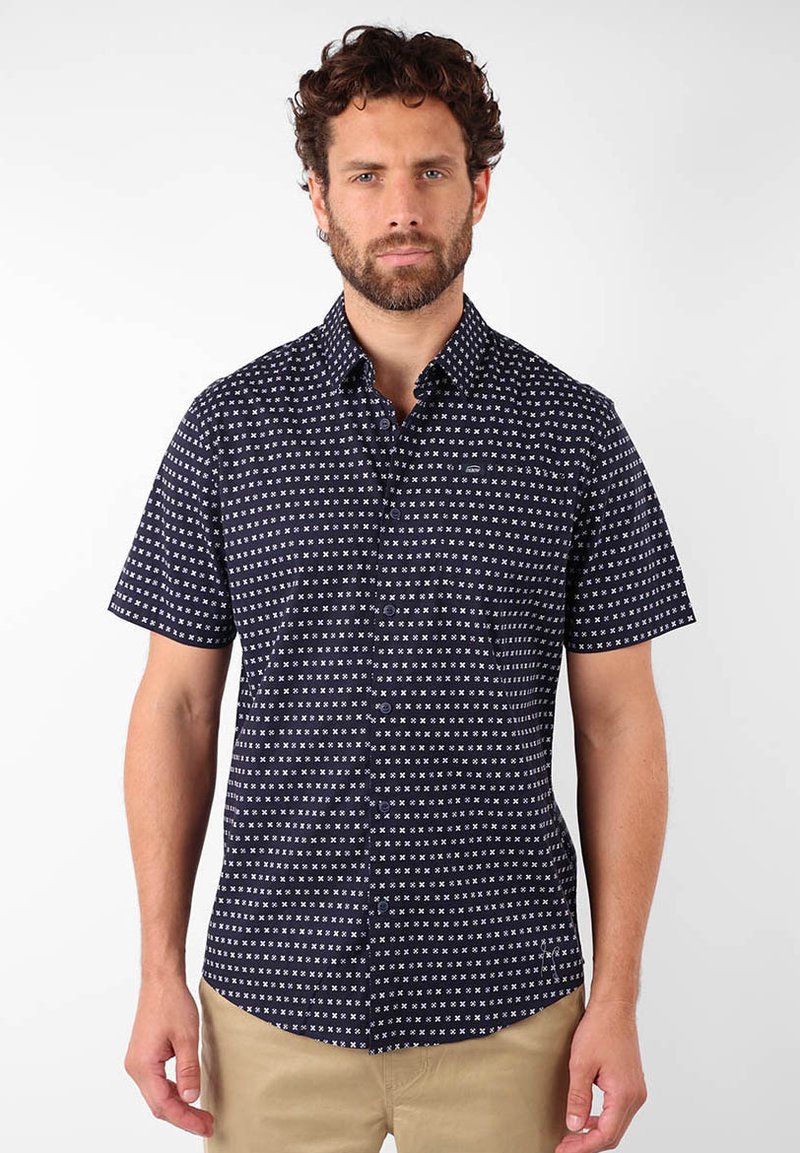 Chemise à manches courtes, de couleur marine, avec un motif géométrique blanc, un col et une poche frontale ; fabriquée en tissu léger.