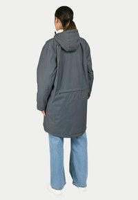 Doudoune grise longue avec capuche, dotée d'une fente au dos et d'une texture lisse, associée à un jean bleu clair à jambes larges et des baskets blanches.