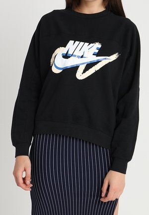 Person iført en sort Nike sweatshirt med en blå og beige logo, kombineret med et mørkeblå skørt med hvide lodrette striber og en sideslids.