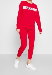 Sudadera roja con una banda horizontal blanca que presenta el logo "HUGO"; combinada con pantalones rojos ajustados y zapatillas blancas. Textura suave en toda la prenda.