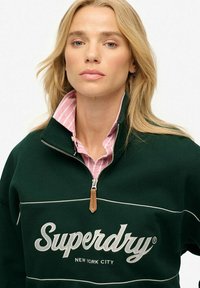 Groene rits-hoodie met witte logo "Superdry" en "New York City" in cursief schrift. Gelaagd over de kraag van een roze gestreept shirt.