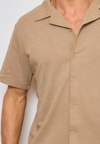 Chemise à manches courtes beige en tissu côtelé, dotée d'un col et de six boutons, avec une surface texturée et une coupe décontractée.
