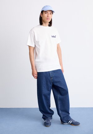 White cotton t-shirt with blue lettering, paired with loose-fitting blue denim jeans and blue cap, on a model standing on blue flooring.