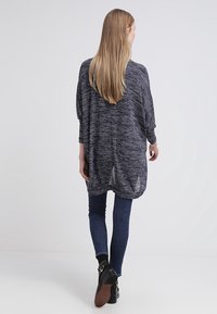 Top leggero a maniche lunghe di colore blu navy con una texture melangiata, caratterizzato da una vestibilità ampia e spacchi laterali. Indossato sopra jeans in denim scuri e stivaletti.