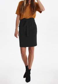 Jupe crayon noire avec une ceinture à nouer, dotée d'une taille haute, d'un tissu structuré et d'une texture lisse, associée à un t-shirt moutarde et des bottines.