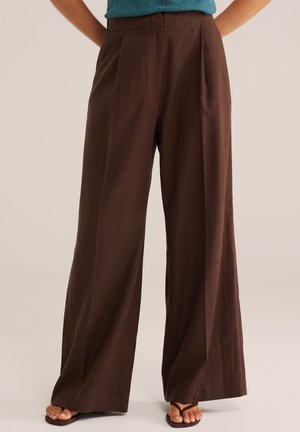 Barna, széles szárú lenvászon nadrág türkizkék felsővel és fekete papucs szandállal viselve, kezek a hát mögött pihennek, semleges háttér előtt elválasztva.