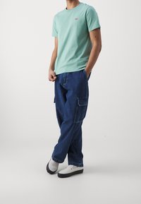 T-shirt en coton vert menthe avec un petit logo rouge, associé à un pantalon cargo en denim bleu ample avec des poches latérales et une finition texturée.