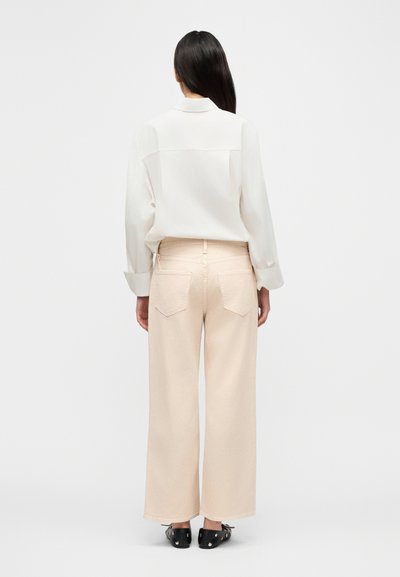 Γυναίκα με μακριά σκούρα μαλλιά, γυρισμένη με την πλάτη, φοράει χαλαρό λευκό πουκάμισο, ανοιχτό μπεζ παντελόνι με φαρδιά πόδια και μαύρα παπούτσια με τρουκς σε απλό φόντο.