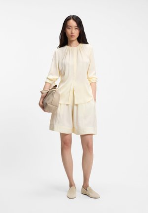 Femme portant un chemisier crème à manches longues et un short assorti, tenant un sac à main tissé beige et portant des chaussures crème sans lacets.