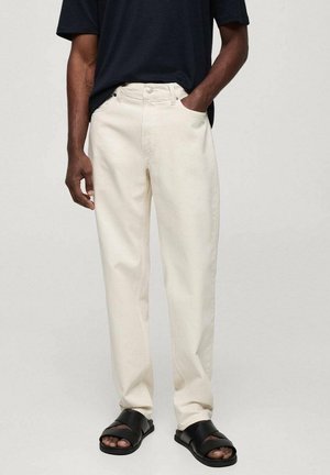 Persona che indossa pantaloni dritti bianco sporco, camicia nera a maniche corte e sandali neri con punta aperta, in piedi davanti a uno sfondo semplice.