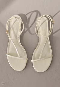 Sandali piatti con cinturini di colore bianco sporco, realizzati in un materiale sintetico liscio, con sottili cinghie alla caviglia e un design minimalista con suola piatta.