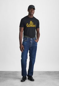 Black short-sleeve t-shirt with "Mr. Catenacci" printed in yellow, paired with dark blue denim jeans and black shoes. Additional black cap worn.