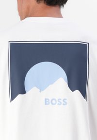Baltas kokvilnas t-krekls ar kalnu un zila apļa grafiku. Vārds "BOSS" izdrukāts gaismas zilā krāsā zem kalna silueta.