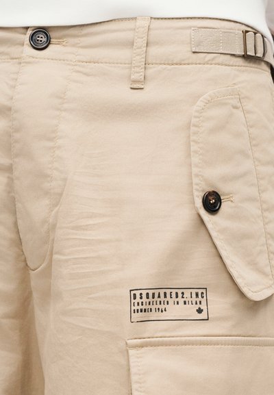 Μπεζ σορτς με λεία υφή, διαθέτουν εμπρόσθιο κλείσιμο με κουμπί, τσέπη με καπάκι και ετικέτα που γράφει "DSQUARED2" σε μαύρο χρώμα.