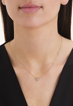 Primer plano de una persona que lleva un delicado collar de cadena de oro con un pequeño colgante de diamante en forma de corazón, sobre una camiseta negra.