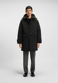 Uomo che indossa un cappotto nero con cappuccio e chiusura a toggle, pantaloni grigi e scarpe nere in piedi contro uno sfondo semplice.