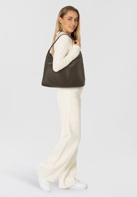 Dunkelgraue Leder-Schultertasche mit strukturierter Oberfläche, abgerundeter Form und langen Riemen, gehalten von einem Modell in einem weißen Outfit.