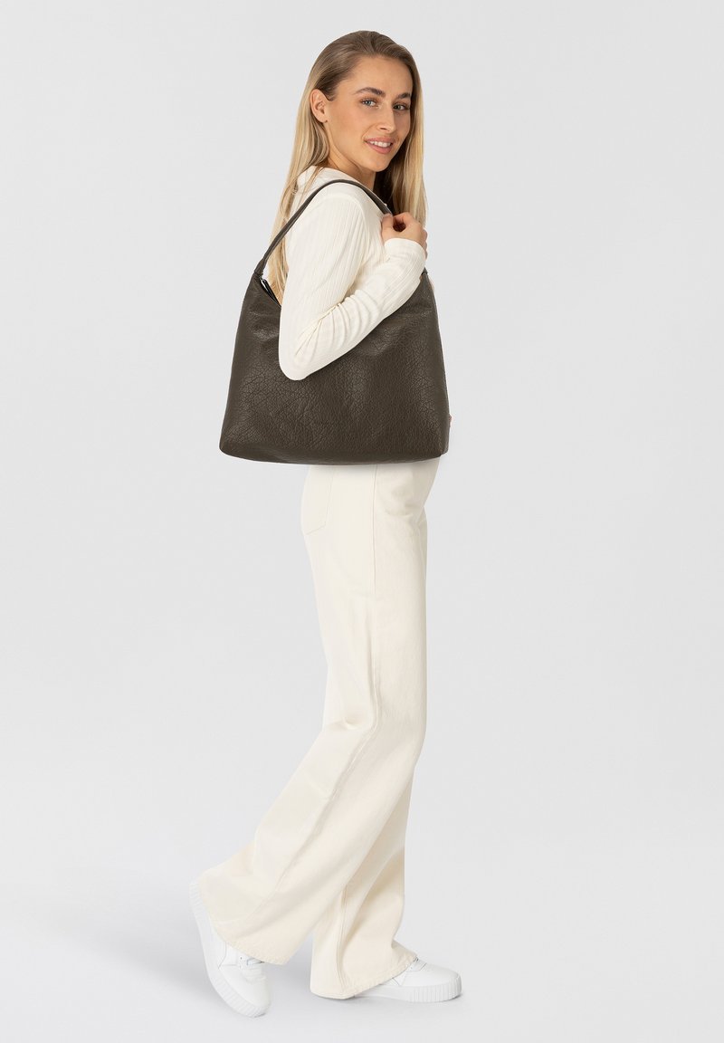 Dunkelgraue Leder-Schultertasche mit strukturierter Oberfläche, abgerundeter Form und langen Riemen, gehalten von einem Modell in einem weißen Outfit.