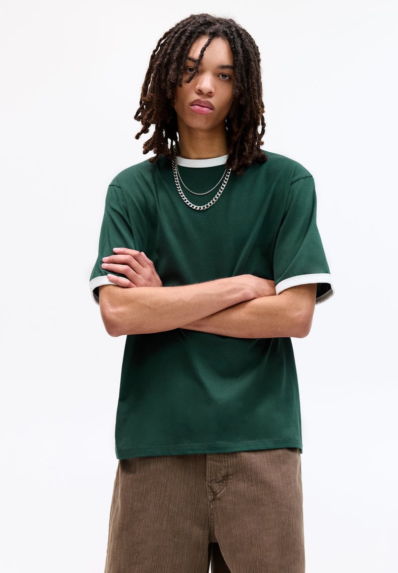 Grøn kortærmet t-shirt med en hvid rund halsudskæring og kontrastkant. Modellen bærer en sølv kædehalssmykke, samt brune fløjl bukser.