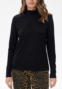 Kobieta ubrana w czarny, długi rękaw z teksturowanego materiału i spodnie w panterkę, stojąca na tle białej ściany.