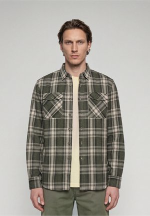 Man met kort bruin haar die een groen en beige geruit knoopoverhemd draagt over een lichtgele T-shirt tegen een grijze achtergrond.
