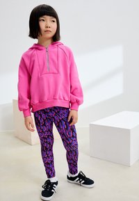 Fuchsiafarbener Hoodie mit halbem Reißverschluss, kombiniert mit lila Leggings mit lebendigem abstraktem Muster, abgerundet durch schwarze Turnschuhe.