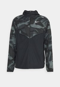 Zwarte jas met een geometrisch camouflagedessin in grijs- en groentinten. Heeft een ritssluiting en een hoge kraag. Gemaakt van lichtgewicht materiaal.