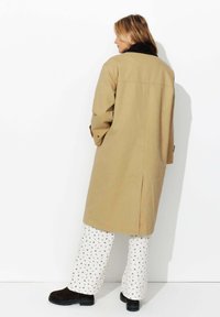 Manteau long beige avec un col marron, fentes latérales et poignets boutonnés, associé à un pantalon blanc à petit motif floral et des bottes noires.