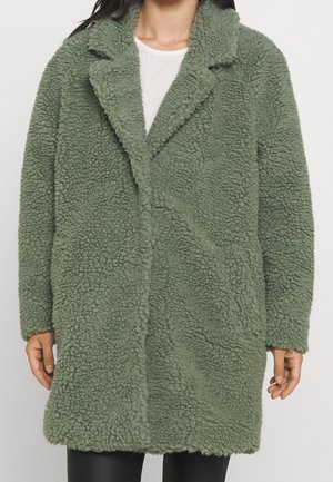 Femme portant un manteau teddy vert texturé oversize par-dessus une chemise blanche et un pantalon noir, debout devant un fond uni.