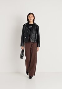 Μαύρο δερμάτινο μπουφάν μπουφάν με ασημένια φερμουάρ, συνδυασμένο με καφέ παντελόνια φαρδιάς γραμμής. Το μοντέλο κρατά μια τσάντα με πουά μοτίβο.