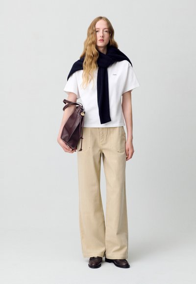 Camiseta blanca de algodón con logo, bufanda azul marino colgada sobre los hombros, pantalones de pierna ancha beige con bolsillos laterales y bolso de cuero ciruela.