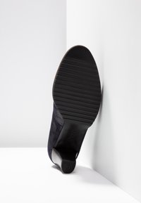 Chaussure en daim bleu marine avec un talon noir texturé et haut, et une semelle en caoutchouc noire striée ; positionnée en angle sur un fond blanc.