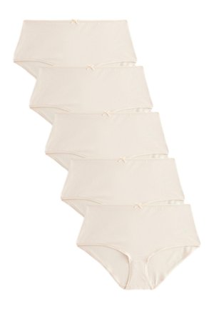 Vrouw draagt beige naadloos hooggetailleerd ondergoed met klein strikdetail, staand met handen ontspannen langs haar zij.