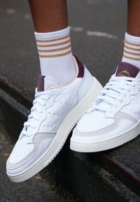 Vita lädersneakers med ljusgrå mockadetaljer, en tjock vit sula och lila hälflik. Bärs tillsammans med vita strumpor med orange ränder.