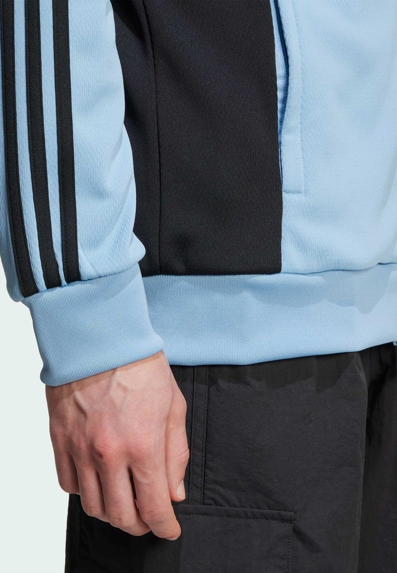 Haut de sport bleu clair avec des accents noirs, présentant trois bandes noires le long de la manche et un poignet côtelé. Texture lisse.