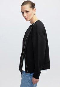 Zwarte sweatshirt met een rits aan de voorkant en een oversized ontwerp. Gemaakt van zachte stof met geribde manchetten. Gecombineerd met blauwe jeans en een witte top.