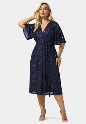 Femme portant une robe portefeuille bleu marine à sequins avec des manches flottantes et des sandales argentées à talons hauts, posant avec une main sur la tête.