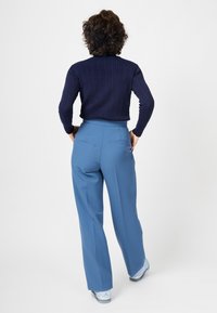 Pantalons larges bleus avec une texture lisse, taille haute et poche arrière, associés à un pull côtelé bleu marine. Présentés de dos.