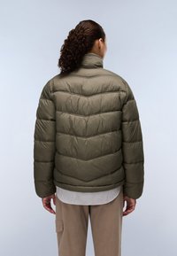 Olijfgroene pufferjack met een chevronpatroon, hoge kraag en ritssluiting. Draagt over een crèmekleurig gestreept shirt en beige broek.