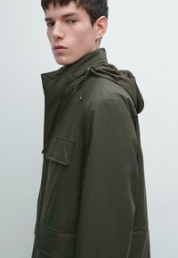 Parka verde oliva con chiusura frontale a zip, cappuccio regolabile e tasche laterali. Il tessuto sembra liscio con una finitura opaca.