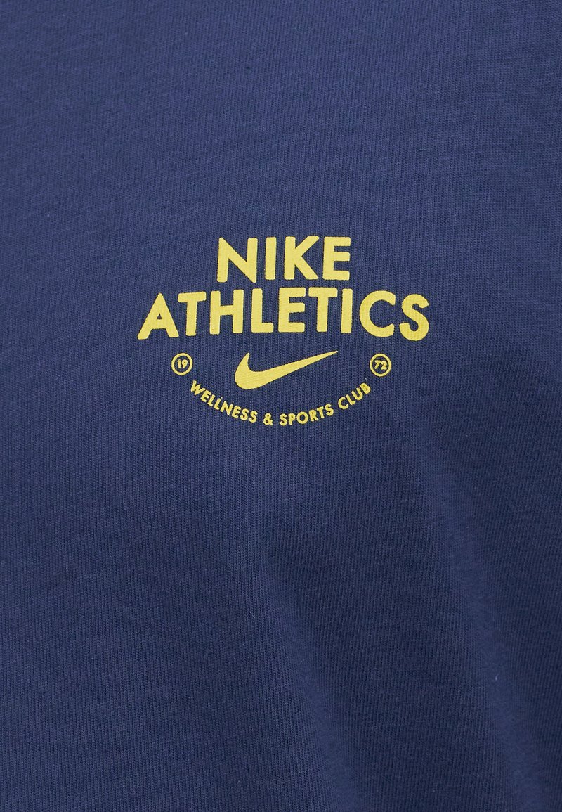 Logo giallo "Nike Athletics Wellness & Sports Club 1972" con il simbolo Nike su sfondo di tessuto blu scuro.