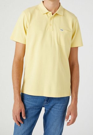Mann trägt hellgelbes kurzärmeliges Poloshirt mit Brusttasche und blaue Jeans, steht mit entspannten Armen vor einfarbigem Hintergrund.