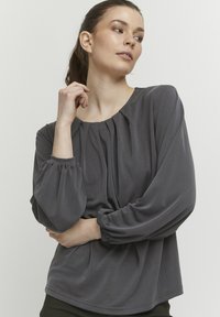 Mørkegrå bluse lavet af glat stof, med rund halsudskæring, plisseringer foran og blussede lange ærmer med elastiske mansjetter.